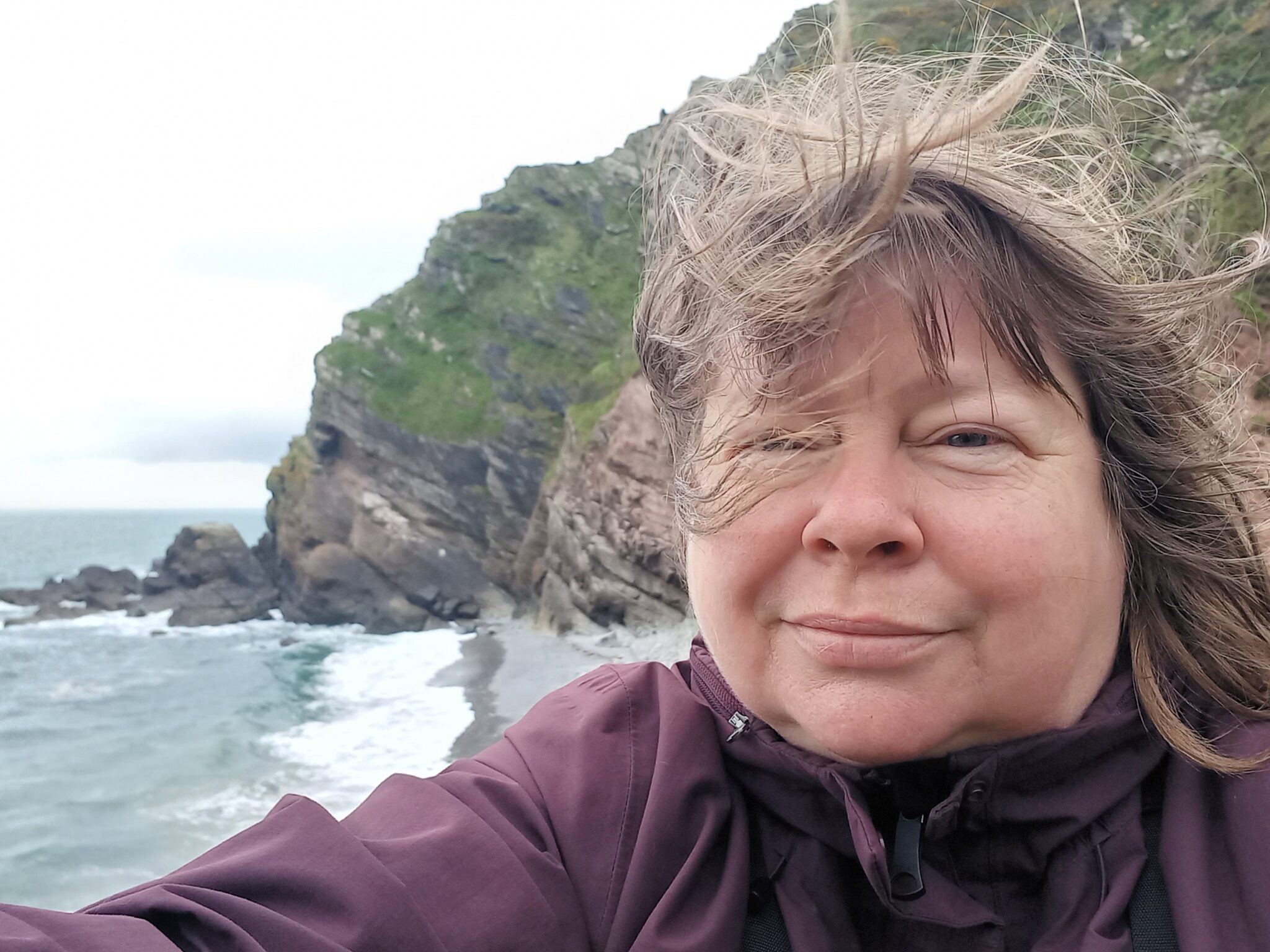 A white person taking a selfie with sea and coast behind them. It is very windy, so their hair is flying around and obscuring part of their face.