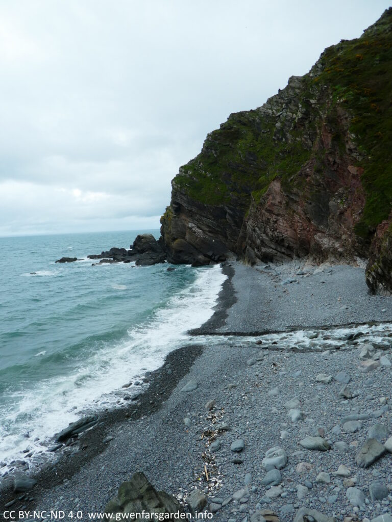 A rocky grey stone beach with some steep cliffs and waves breaking on the shoreline.