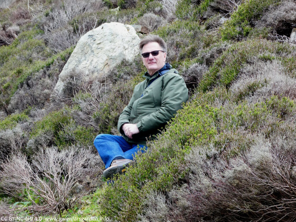 A white person sitting amongst the heather and rocks on the side of a hill.