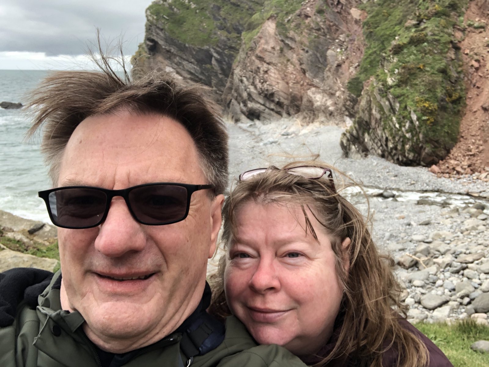 A selfie of two white people looking very happy, with the sea and coast behind them.