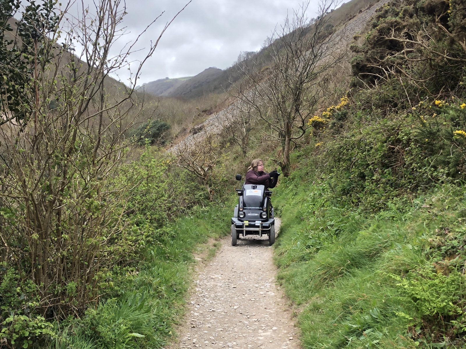 The white person on the tramper, only they aren't looking at the camera, because they are taking photos of wildflowers! You can see hints of high hills & a steep valley, behind.